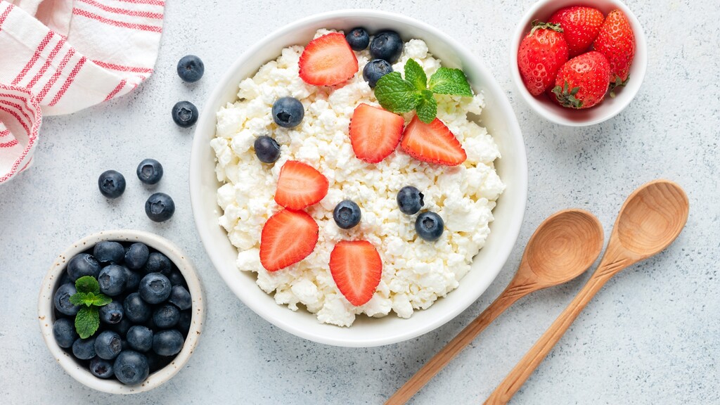 Cottage cheese topped with strawberries, blueberries, and mint, served with fresh berries and wooden spoons on a light kitchen surface.