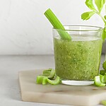 Glass of blended celery juice with fresh celery stalks, chopped celery, and a lemon wedge on a kitchen cutting board.