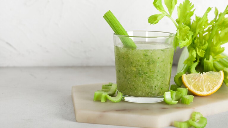 Glass of blended celery juice with fresh celery stalks, chopped celery, and a lemon wedge on a kitchen cutting board.