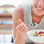 11 Natural Ways to Lower Cortisol and Calm Your System Woman eating a breakfast bowl with berries, oats, and yogurt in a bright kitchen.
