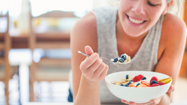 Woman eating a breakfast bowl with berries, oats, and yogurt in a bright kitchen.