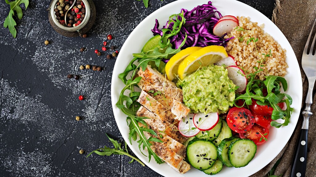 Grilled chicken bowl with quinoa, avocado, cucumber, tomatoes, cabbage, greens, and lemon on a dark table.