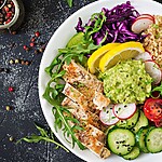 Grilled chicken bowl with quinoa, avocado, cucumber, tomatoes, cabbage, greens, and lemon on a dark table.