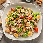 Weight Loss Without Dieting: How to Keep Your Favorite Foods Balanced chicken pasta salad with lettuce, tomatoes, boiled eggs, croutons, and bowtie pasta on a white plate.