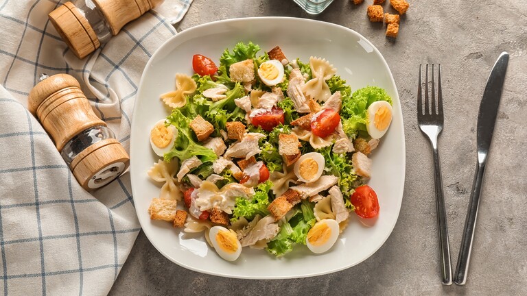 Balanced chicken pasta salad with lettuce, tomatoes, boiled eggs, croutons, and bowtie pasta on a white plate.