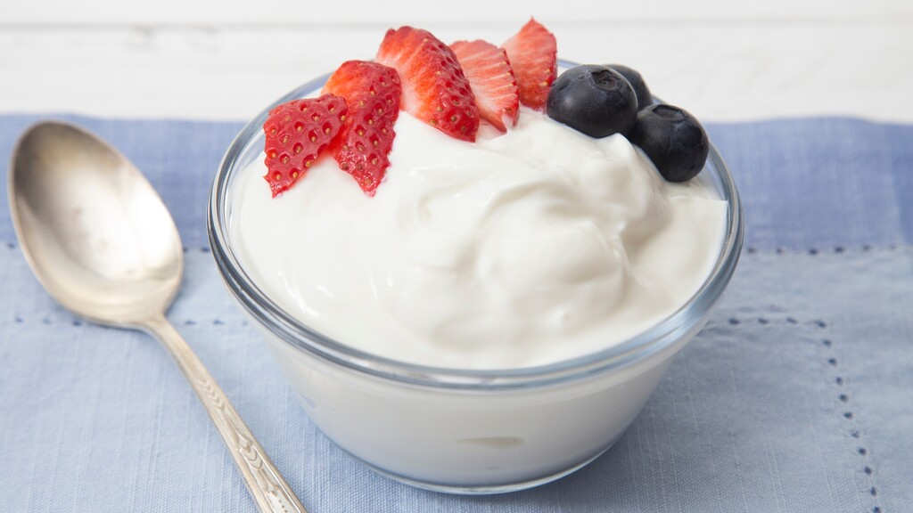 Plain Greek yogurt topped with sliced strawberries and blueberries in a glass bowl beside a spoon on a blue cloth.