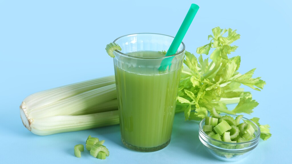 Fresh celery juice in a glass with a straw, surrounded by whole celery stalks, chopped celery pieces, and a small bowl on a blue background.