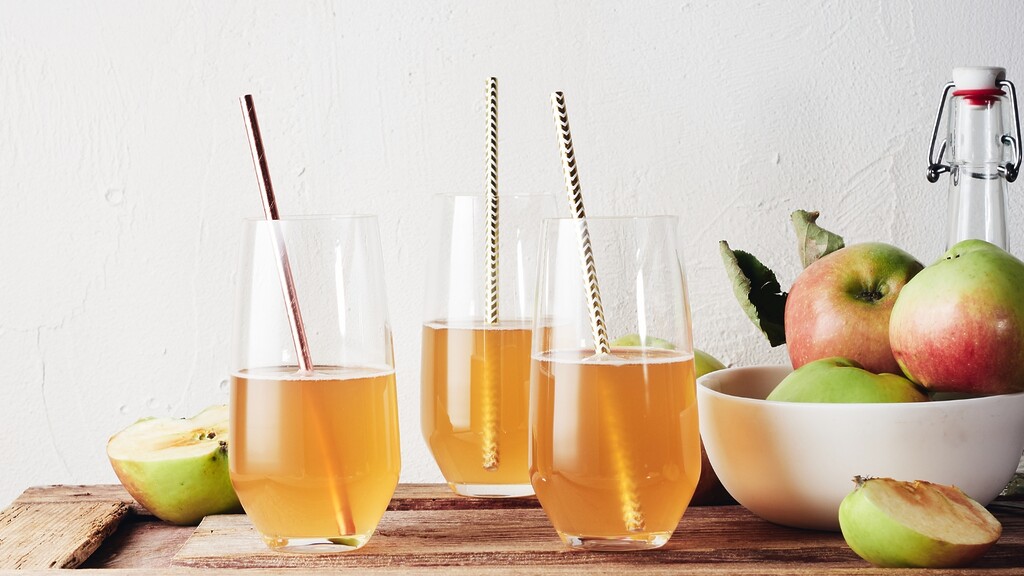 Three glasses of diluted apple cider vinegar drink with straws on a wooden table beside fresh apples and a glass bottle.
