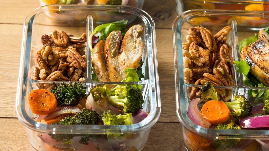 Glass meal prep containers filled with grilled chicken, roasted vegetables, pecans, and leafy greens on a wooden table.