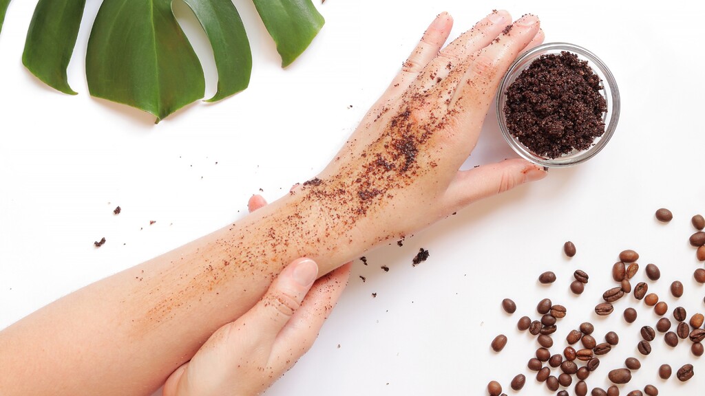 Hands applying a homemade coffee body scrub with coffee grounds, beans, and green leaves on a white surface.