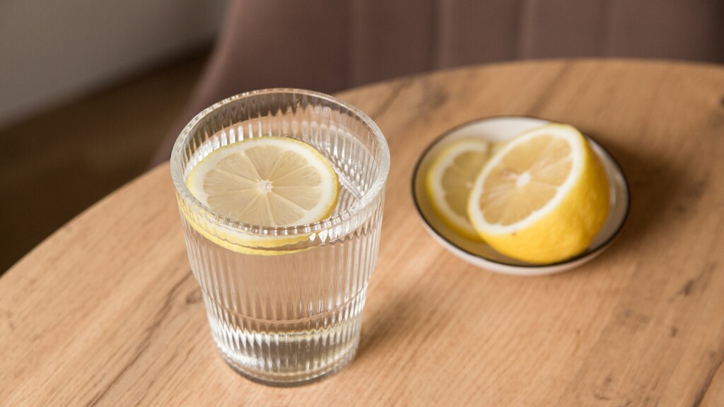 Glass of lemon water on a wooden table with sliced lemons in the background.