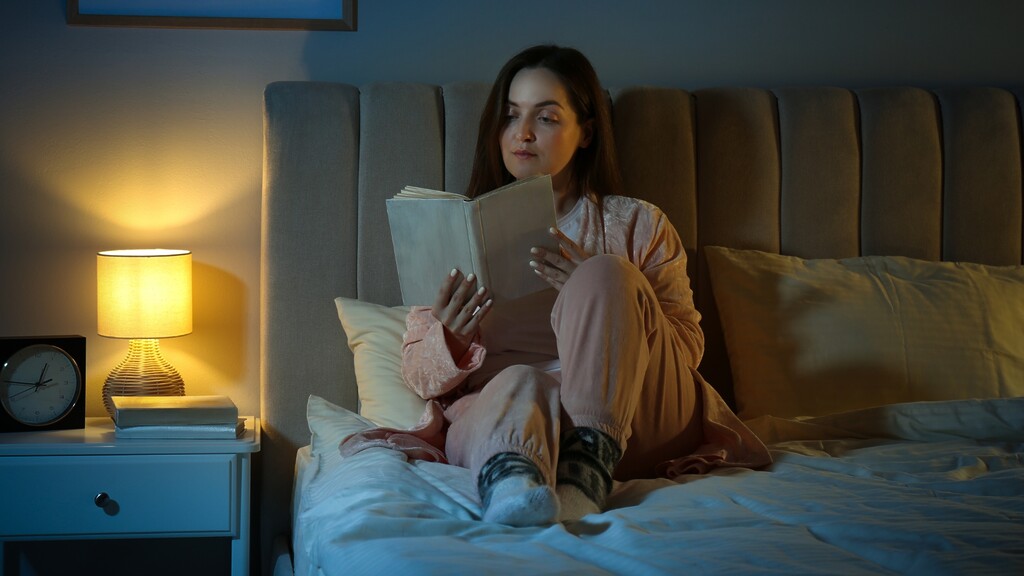 Woman reading in bed at night as part of a relaxing sleep routine for better health.