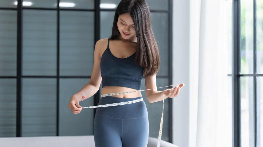Woman measuring her waist with a tape measure while wearing workout clothes in a bright fitness studio.