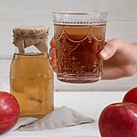 A hand holding a glass of apple cider vinegar drink beside a jar of vinegar and fresh red apples.