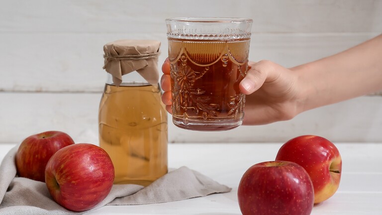 A hand holding a glass of apple cider vinegar drink beside a jar of vinegar and fresh red apples.