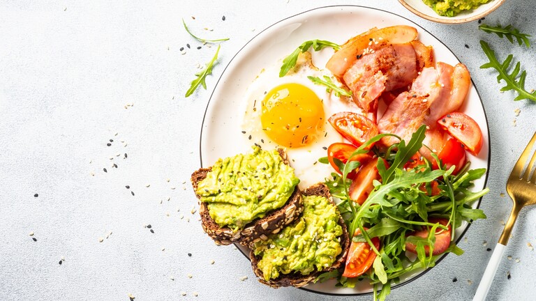 Avocado toast with a fried egg, bacon, tomatoes, and arugula on a breakfast plate.