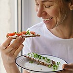 Woman eating a crispbread snack with tomatoes and greens by a window.