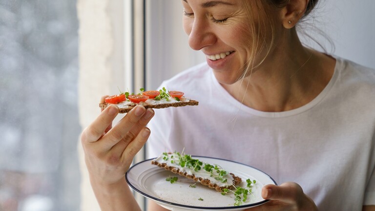 Woman eating a crispbread snack with tomatoes and greens by a window.