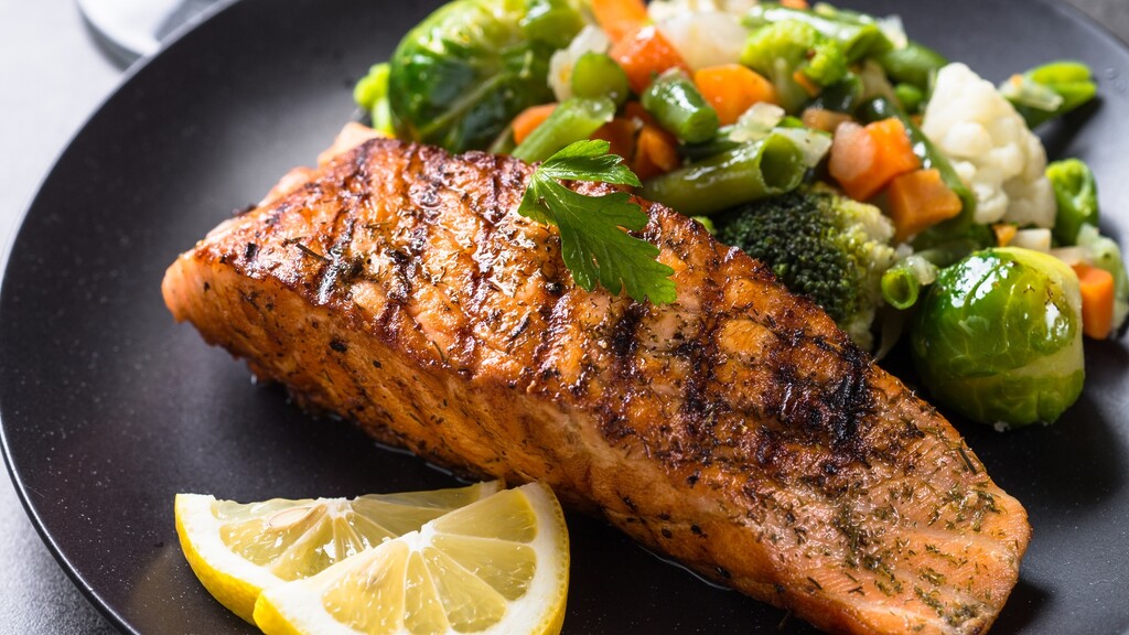 Grilled salmon served with mixed vegetables and lemon on a black plate.