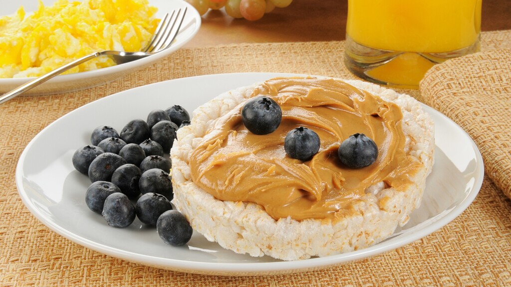 Rice cake topped with peanut butter and blueberries, served with scrambled eggs and orange juice.
