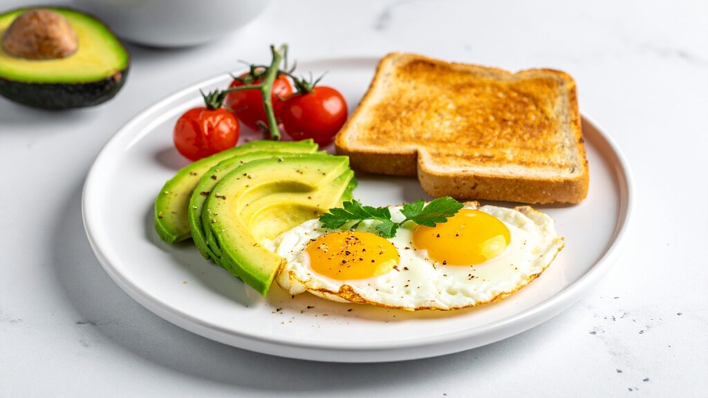 Breakfast plate with fried eggs, sliced avocado, tomatoes, and toast.