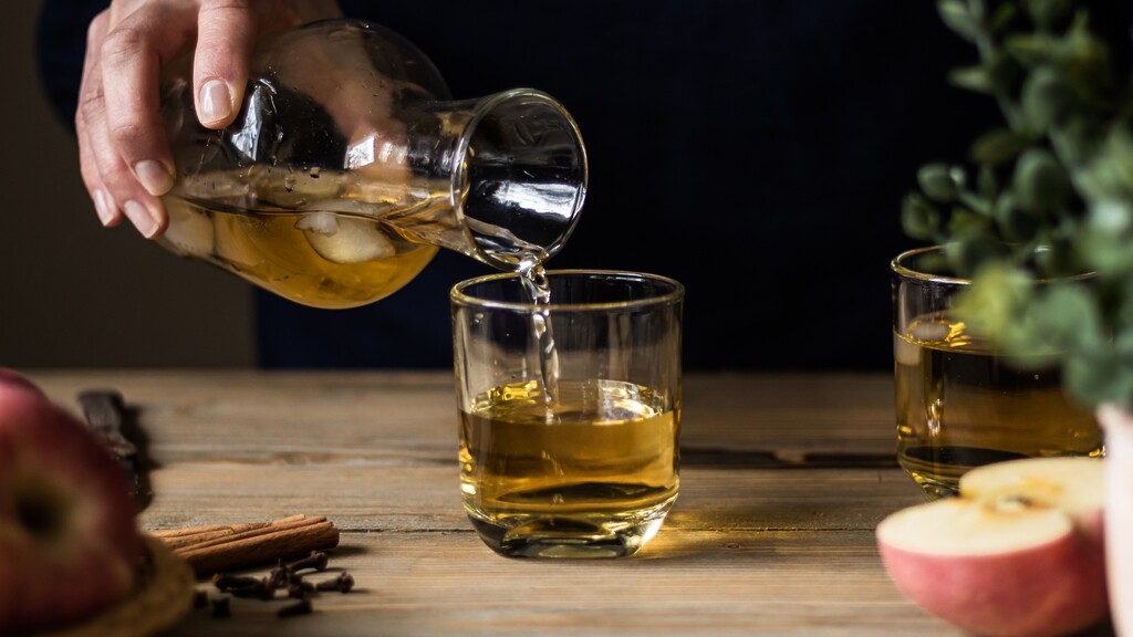 Apple cider vinegar being poured into a glass on a wooden table with apples, cinnamon sticks, and cloves.