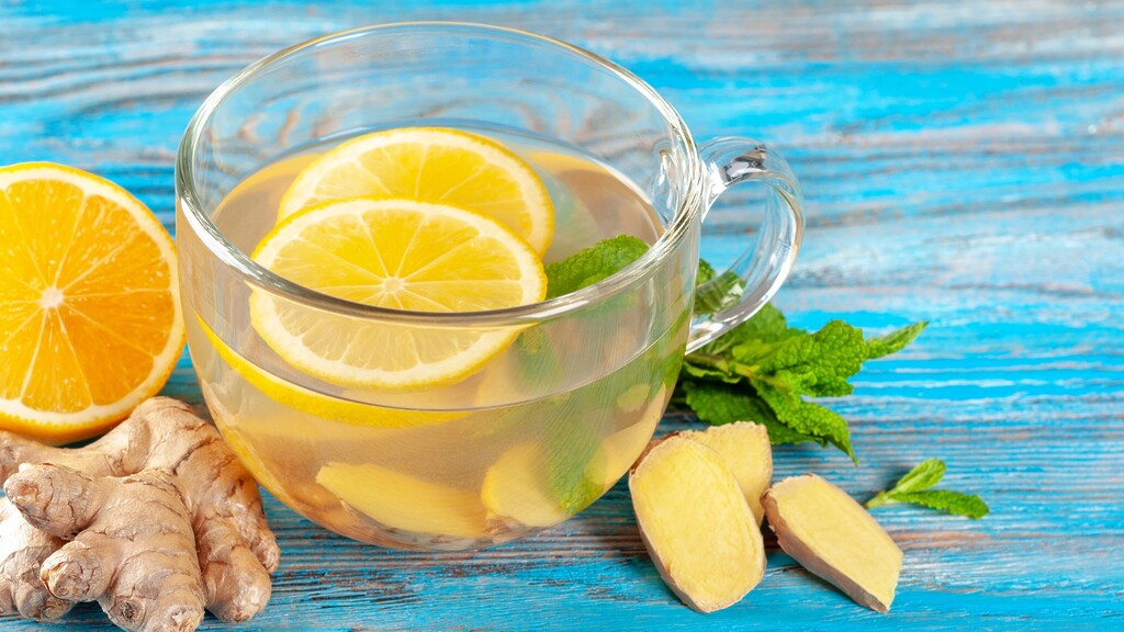Ginger lemon tea in a glass cup with fresh ginger root, lemon slices, and mint on a blue wooden table.
