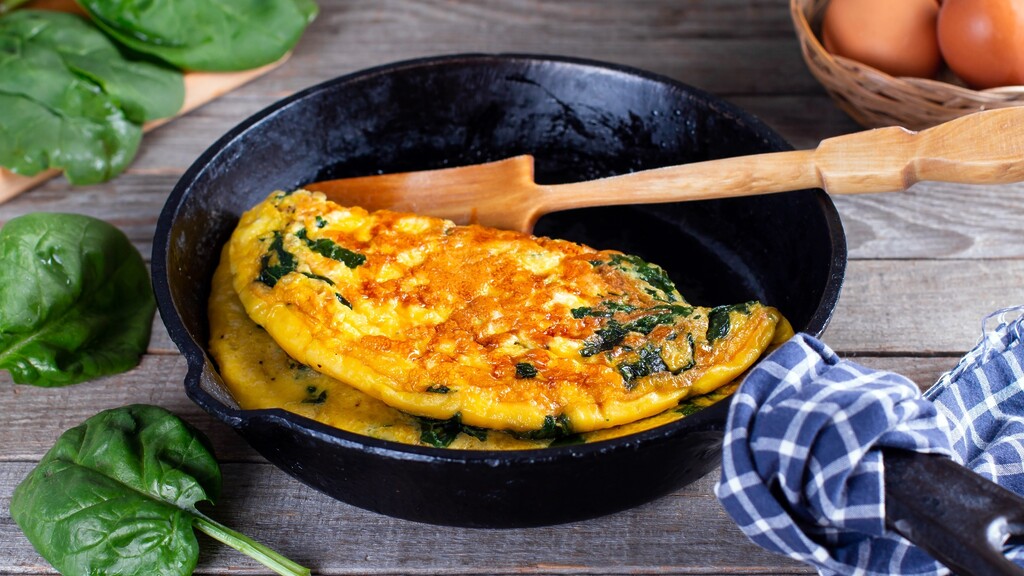 Spinach omelet cooking in a cast iron skillet with fresh spinach and eggs nearby.