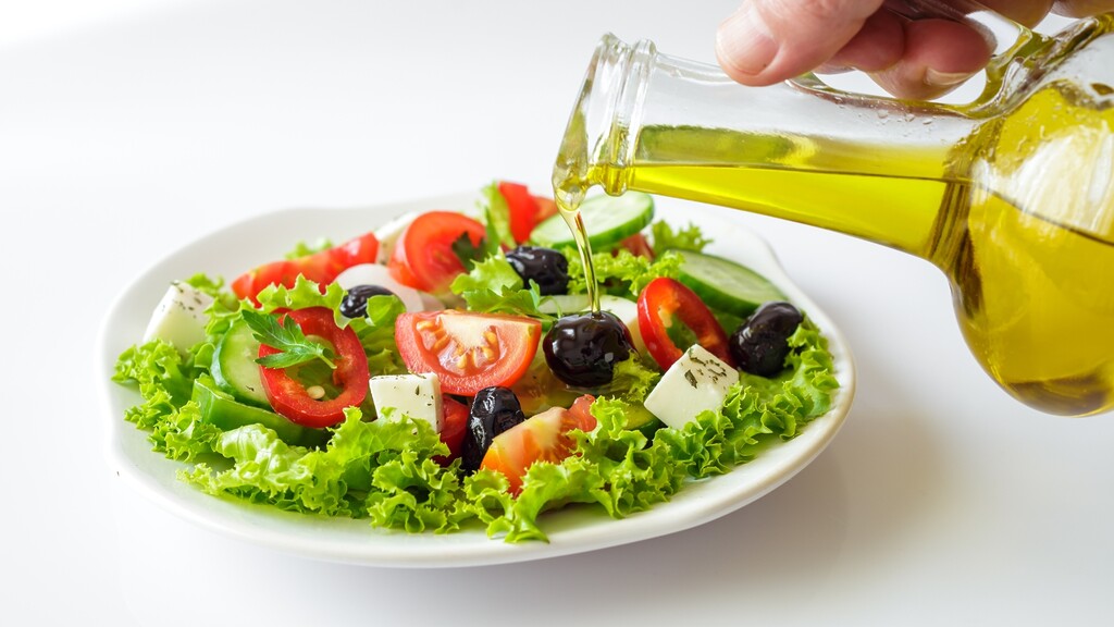 Olive oil being poured over a fresh Mediterranean salad with lettuce, tomatoes, cucumber, olives, and feta.