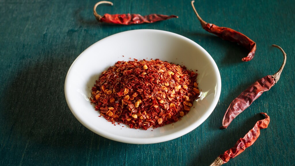 Bowl of crushed red chili flakes with dried chili peppers on a dark table.