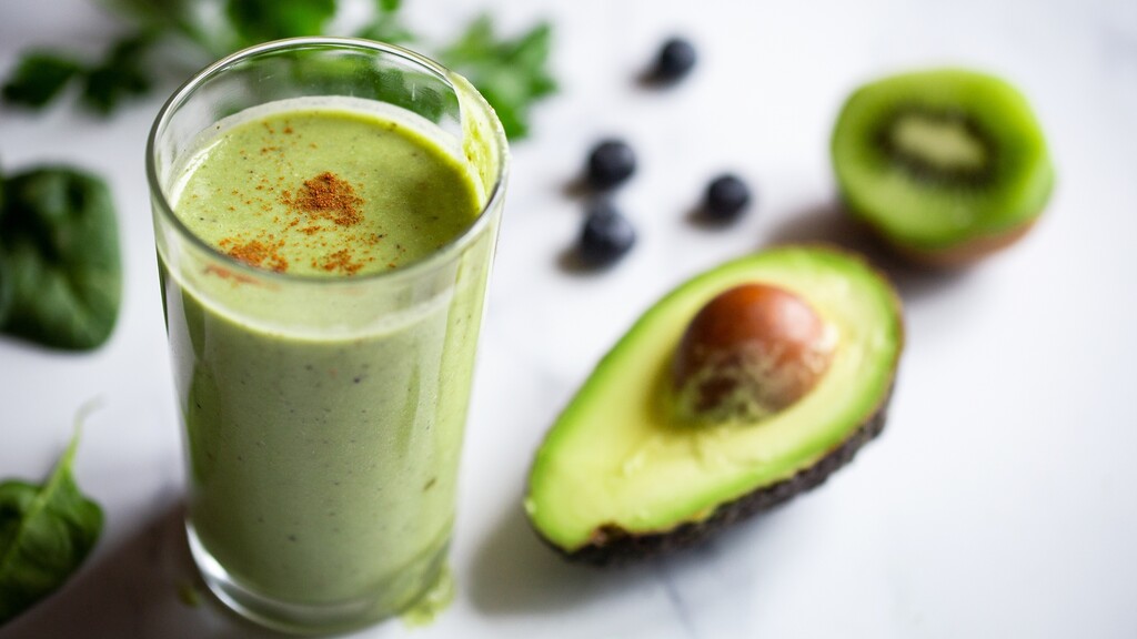 Green avocado smoothie in a glass with spinach, blueberries, and sliced avocado nearby.