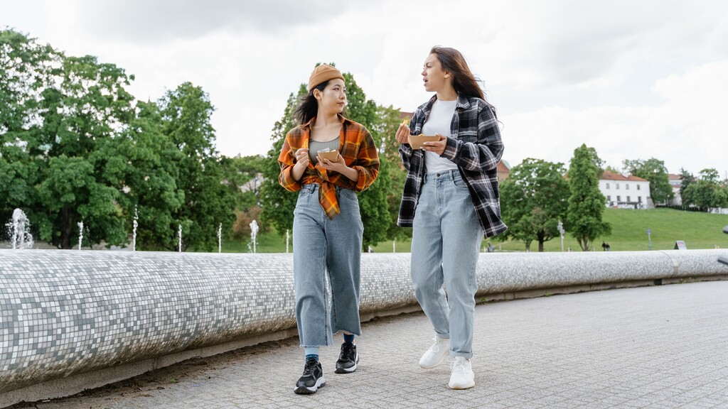 Two women walking outdoors while talking and eating a light snack.