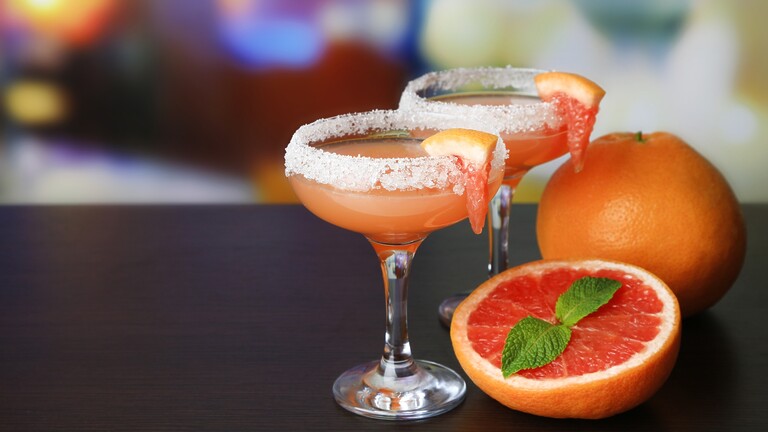 Two grapefruit Paloma cocktails with salted rims, citrus wedges, fresh mint, and whole grapefruit on a bar.