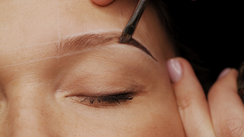 Close-up of a woman’s eyebrow being filled with an angled brush while brow mapping lines guide the shape.