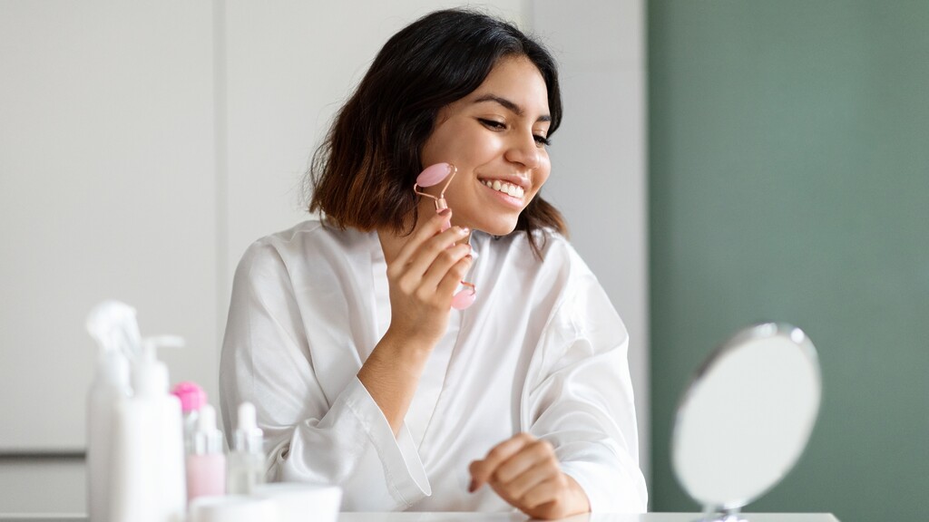 Woman using a pink face roller on her cheek during a morning skincare routine at a vanity mirror.