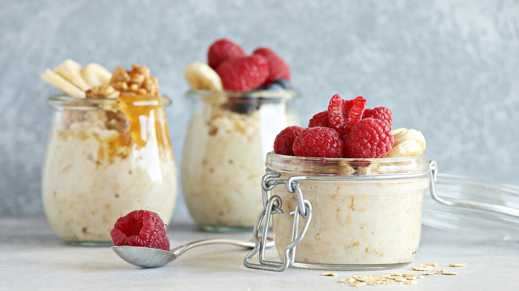 Three jars of overnight oats topped with raspberries, banana, walnuts, and honey on a light kitchen surface.