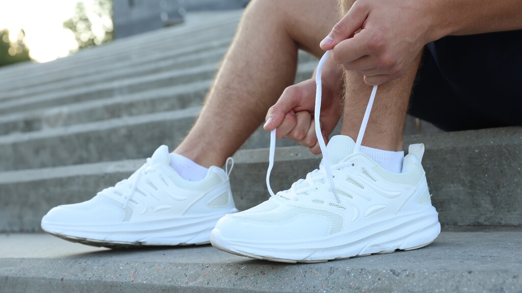 Man tying the laces of white walking shoes while sitting on outdoor steps before a walk.