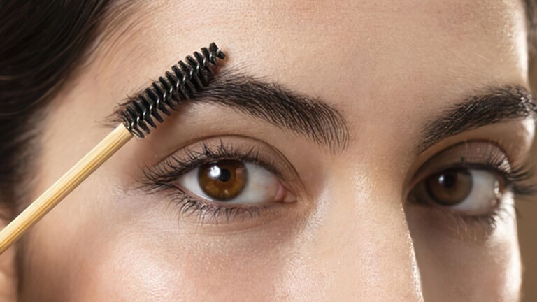 Close-up of a woman brushing a full eyebrow upward with a spoolie for a groomed, natural brow shape.