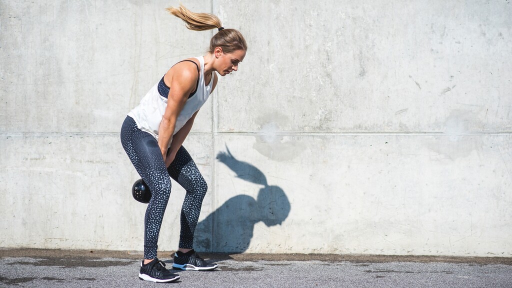 Woman performing a kettlebell swing outdoors against a concrete wall.