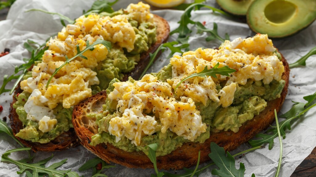 Avocado toast topped with scrambled eggs, black pepper, and arugula on toasted sourdough bread.